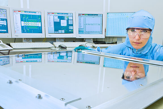 Frau in Laborkleidung in der Hocke vor einem Glas, im Hintergrund Monitore