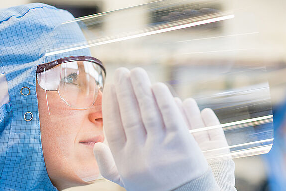 Frau mit blauem Laborkopfschutz, Brille und Handschuhen schaut durch gebogenes Glas