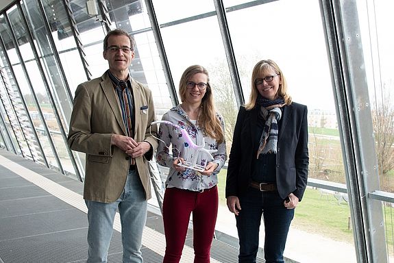 Ein Mann und zwei Frauen stehen vor einem Panoramafenster, sie zeigen das transparente Modell einer Luftröhre mit einem LED-Schlauch darin