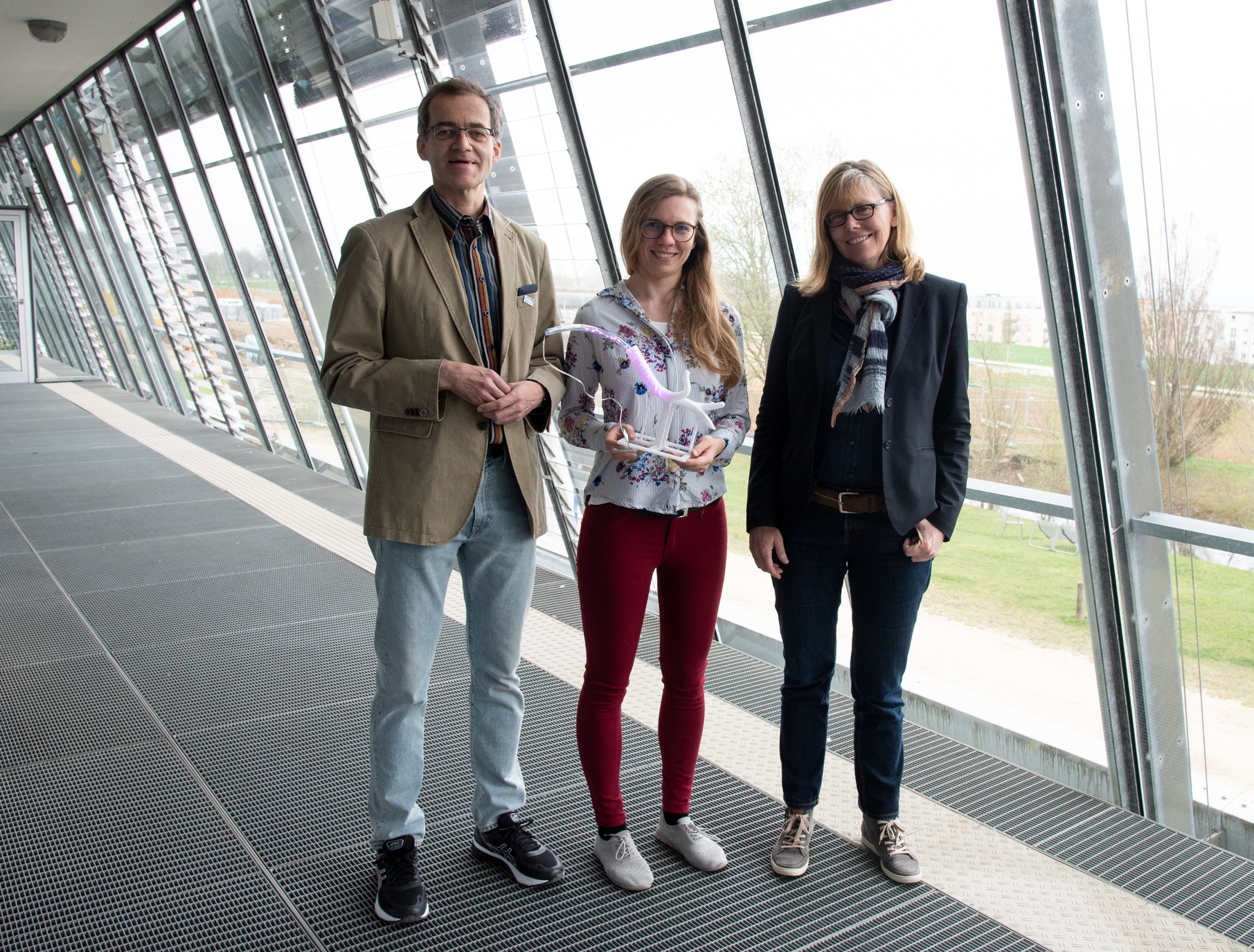 Ein Mann und zwei Frauen stehen vor einem Panoramafenster, sie zeigen das transparente Modell einer Luftröhre mit einem LED-Schlauch darin